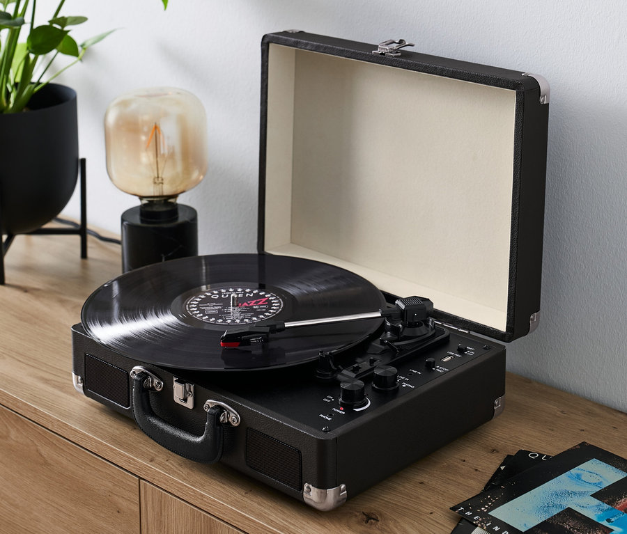 Ein schwarzer, tragbarer Retro-Plattenspieler mit Bluetooth-Funktion spielt eine Schallplatte auf einer Holzkommode ab.