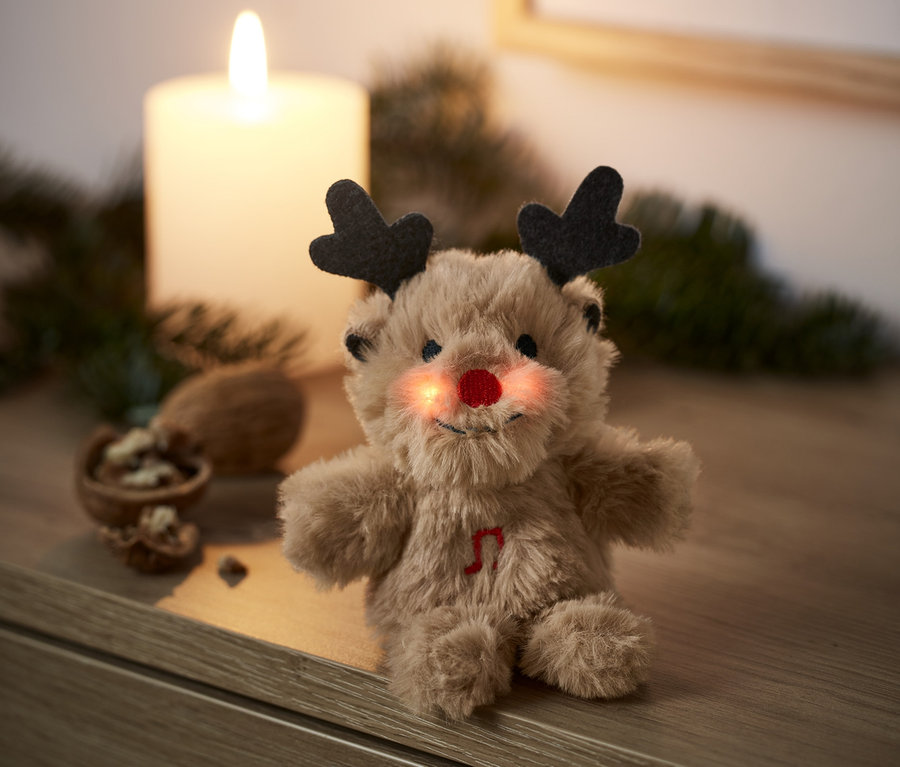 Ein singendes Mini-Plüschtier Rentier mit roter Nase sitzt auf einer Holzkommode neben einer brennenden Kerze.
