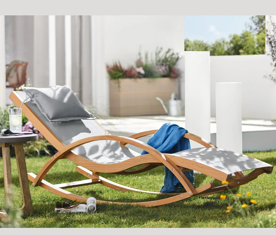 Im Gras stehen eine Teak-Schaukelliege mit Textilen, ein runder Beistelltisch mit Beton-Tischplatte und Solar-Lichtsäulen mit Farbwechsel in verschiedenen Höhen.