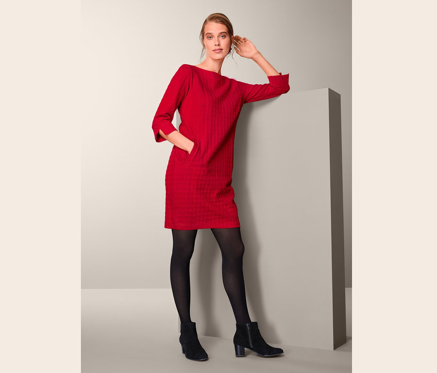 Eine Frau, die sich an eine Wand lehnt, trägt ein rotes Sweatkleid mit 3/4-Arm, eine schwarze Strumpfhose und schwarze Stiefel.