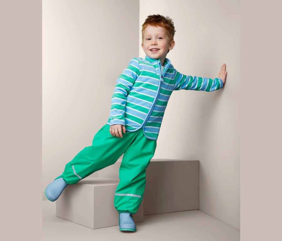 Junge in recycelter Fleecejacke mit blauen Streifen, grüner Hose und blauen Schuhen lehnt an einer Wand.