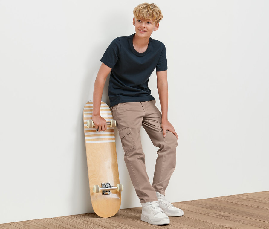 Ein Junge lehnt an einer Wand und hält ein Skateboard. Er trägt eine Kinder-Cargohose.