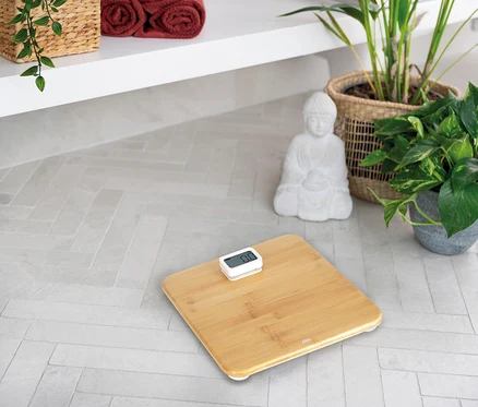 ADE Digitale Bambus-Personenwaage auf hellem Fliesenboden, im Hintergrund Handtücher, Pflanzen und eine Buddha-Statue.