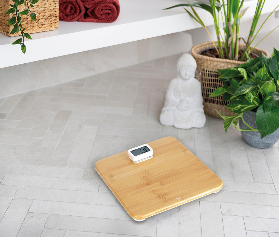 ADE Digitale Bambus-Personenwaage auf hellem Fliesenboden, im Hintergrund Handtücher, Pflanzen und eine Buddha-Statue.