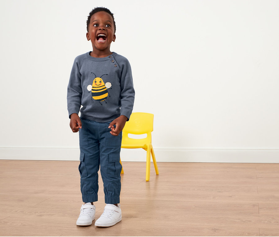 Ein Junge steht und trägt eine blaue Kleinkinder-Schlupfhose und einen Pullover mit einem Bienenbild.