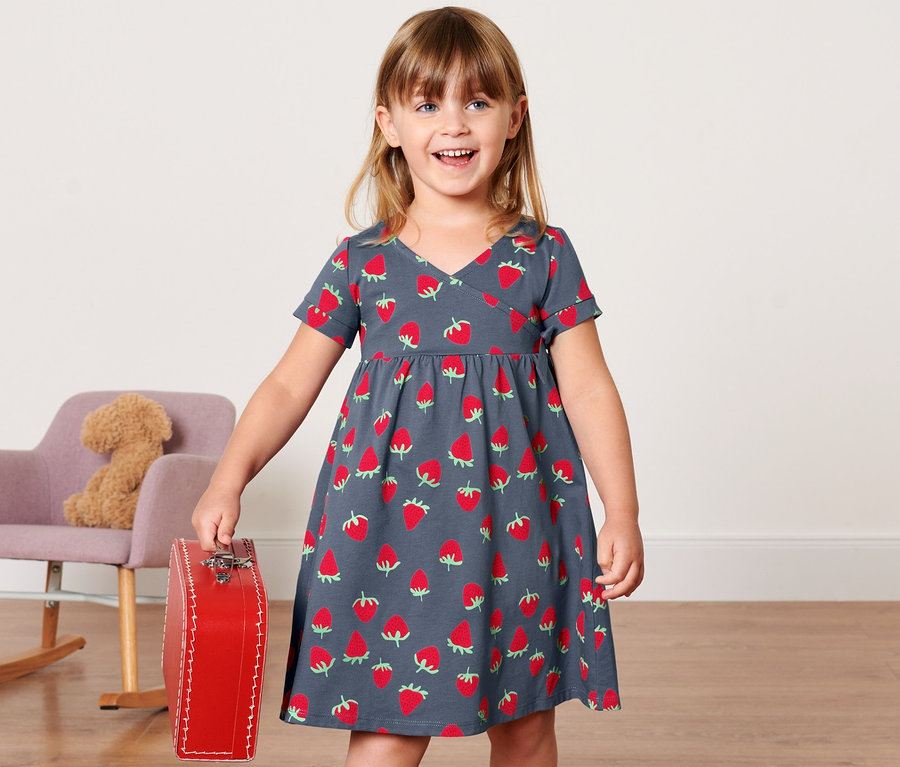 Mädchen hält einen roten Koffer und trägt ein Kleinkinder-Jerseykleid mit Erdbeeren.