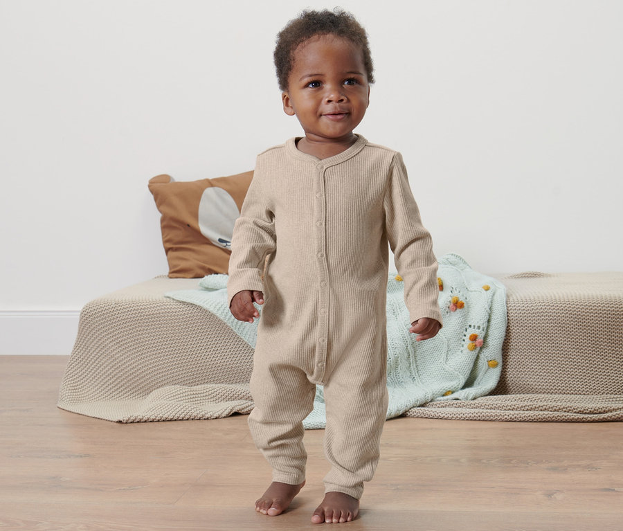 Ein dunkelhäutiges Baby steht auf dem Boden und trägt einen Baby-Strampler.