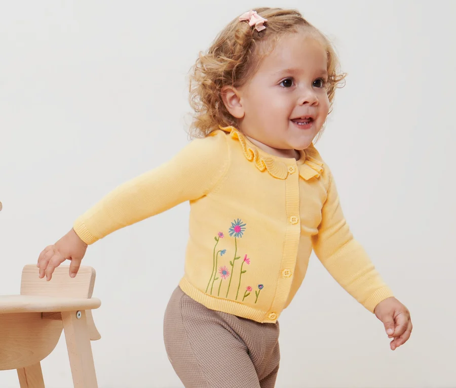 Ein Kind mit einer gelben Baby-Strickjacke steht neben einem hölzernen Schaukelpferd.