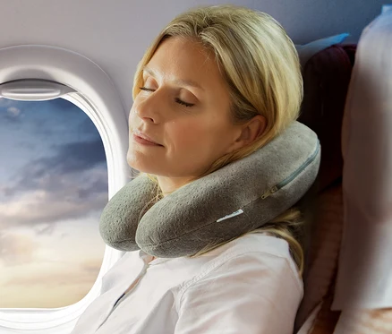 Blonde Frau schläft im Flugzeug mit einem grauen TEMPUR® Transitkissen um den Hals, neben einem Fenster mit Wolken.
