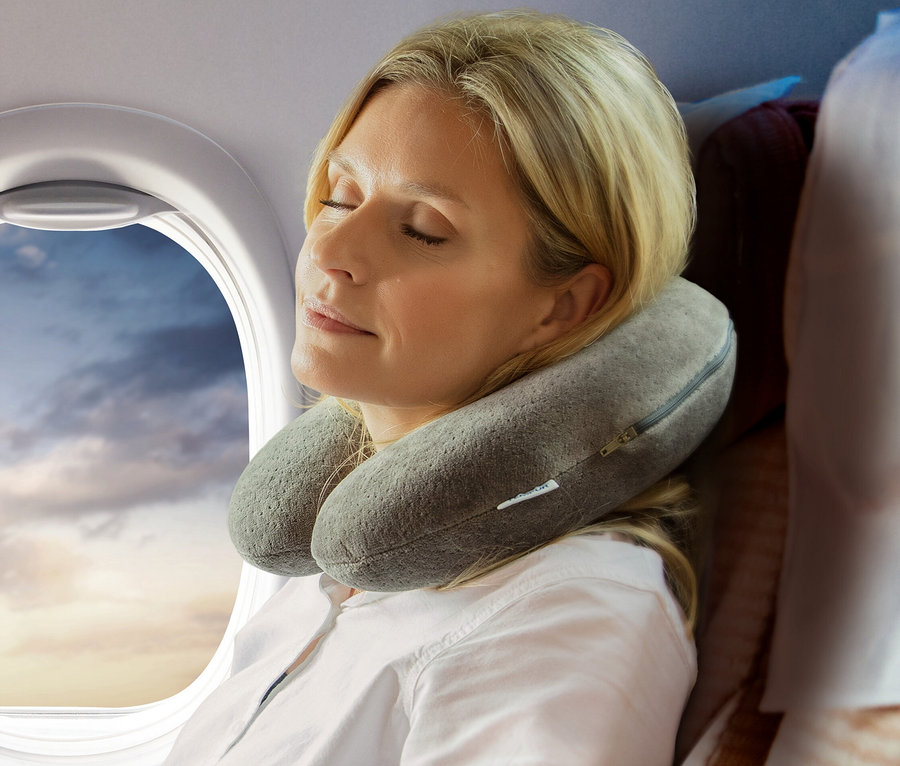 Blonde Frau schläft im Flugzeug mit einem grauen TEMPUR® Transitkissen um den Hals, neben einem Fenster mit Wolken.