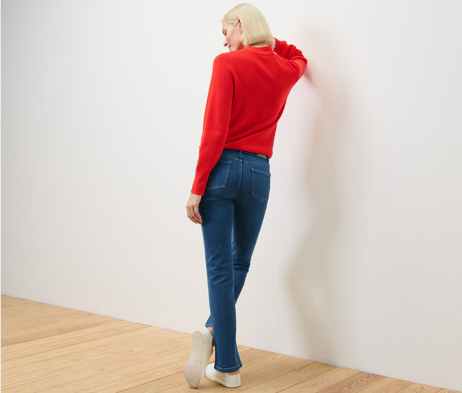 Frau in rotem Pullover und Straight Jeans – Fit »Kira« lehnt an der Wand.