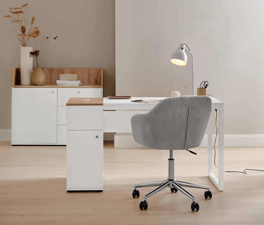 Beiger Schreibtischstuhl steht an einem Schreibtisch mit Stauraum. Im Hintergrund befindet sich ein Sideboard mit Schubladen und Tür und eine Tischleuchte aus Metall.