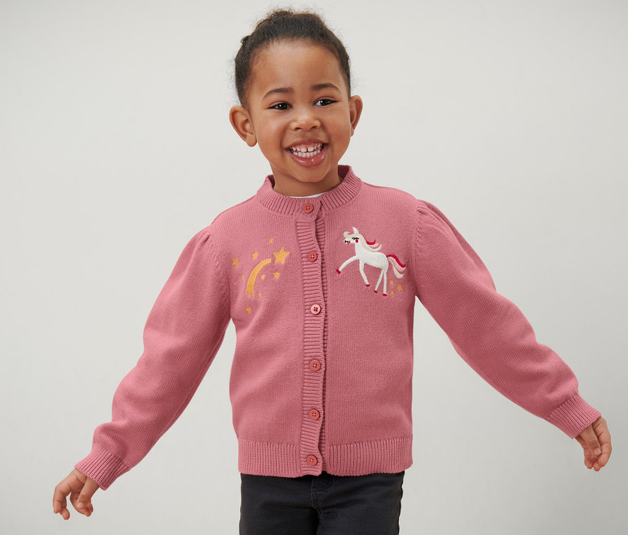 Lächelndes Mädchen trägt eine rosa Kinder-Strickjacke mit Einhorn- und Stern-Applikationen.