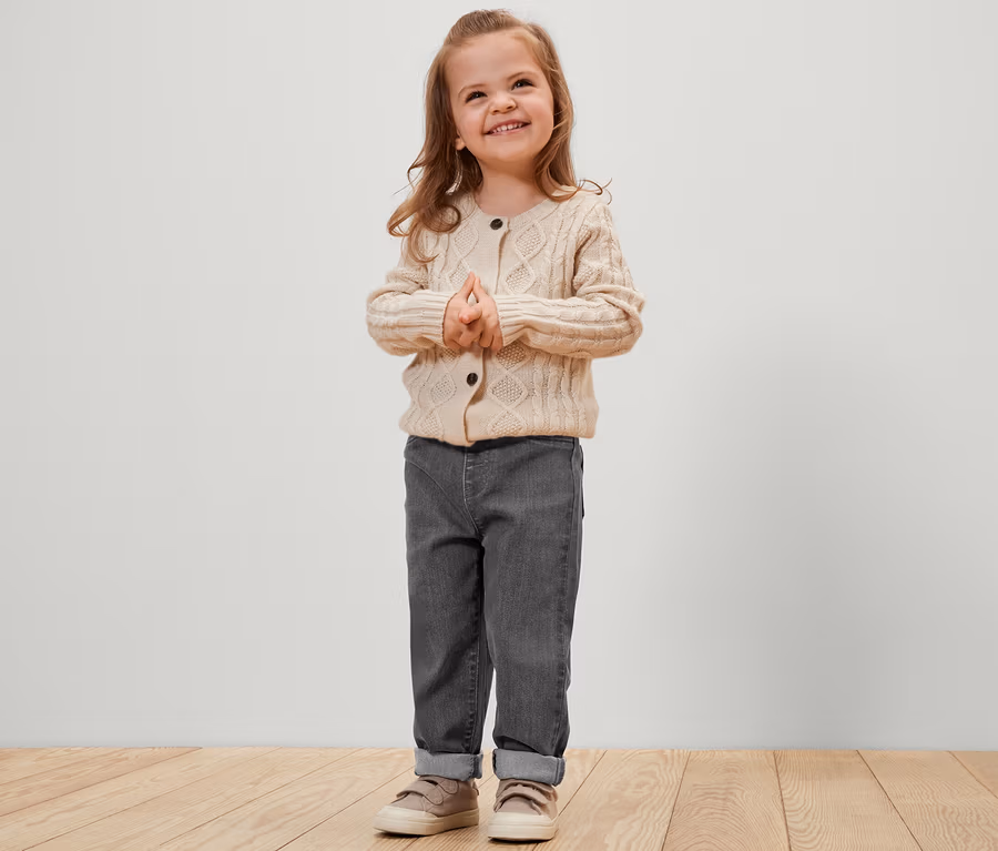 Lächelndes kleines Mädchen steht mit gefalteten Händen da. Sie trägt eine graue Hose und eine cremefarbene Strickjacke. Kinder-Pull-on-Pants – Fit »Luca«, grau.