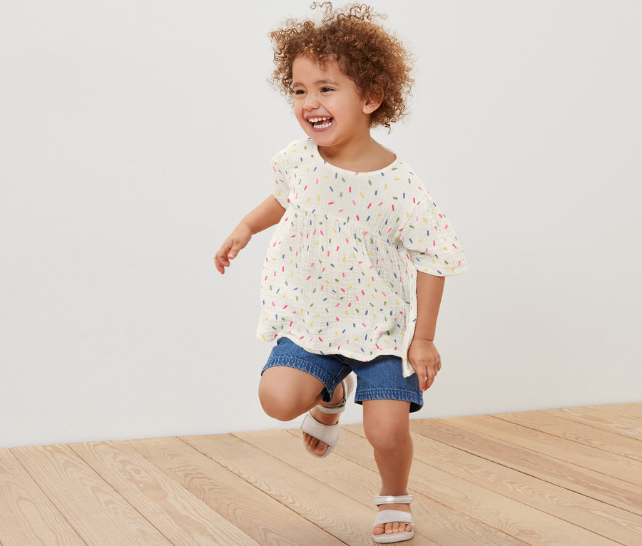 Lächelndes Mädchen mit lockigem Haar rennt und trägt eine Kinder-Musselin-Bluse, Jeansshorts und Sandalen.