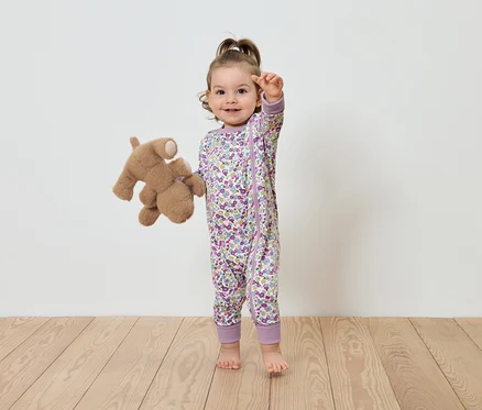 Ein Kind in einem geblümten Overall steht mit einem Plüschteddybär.