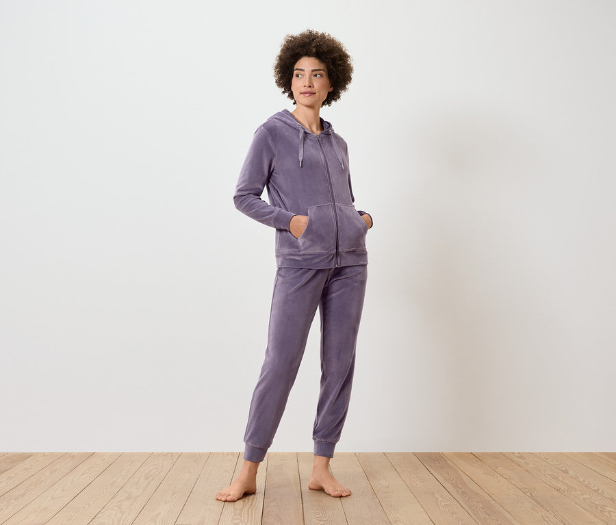 Frau steht und trägt einen lila Trainingsanzug mit Kapuzenpullover und Jogginghose.