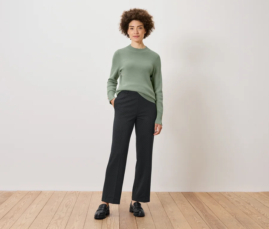 Eine Frau mit lockigem Haar steht mit einer Hand in der Hosentasche. Sie trägt einen grünen Pullover, gemusterte Hosen und schwarze Schuhe.
