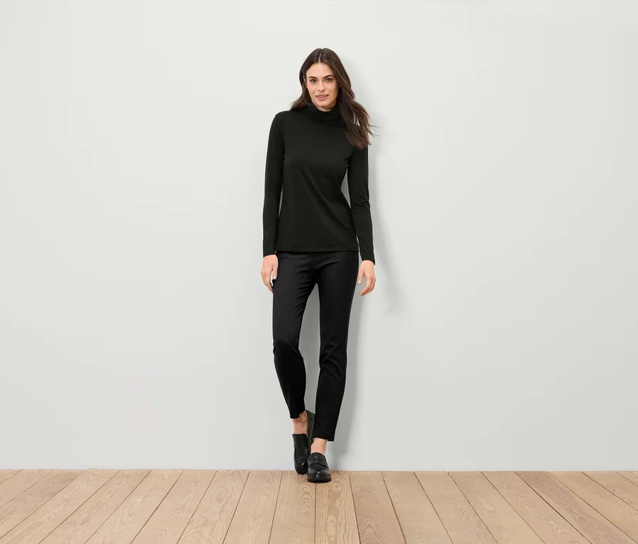 Eine Frau steht und trägt einen schwarzen Rollkragenpullover, eine schwarze Hose und schwarze Schuhe.