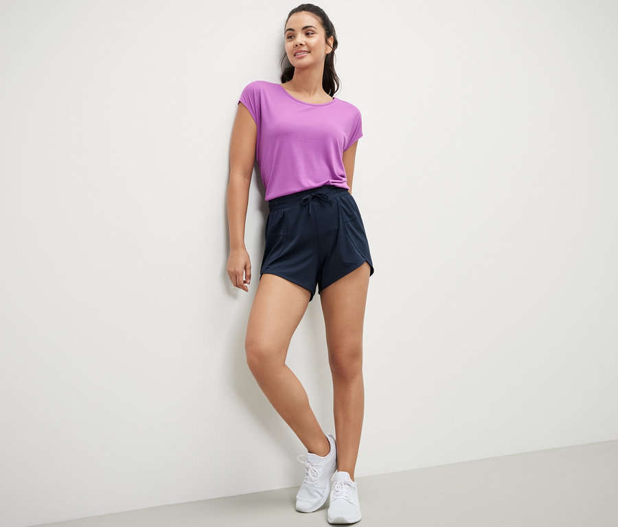 Eine Frau lehnt an einer Wand und trägt ein lila T-Shirt und dunkelblaue Funktionsshorts.