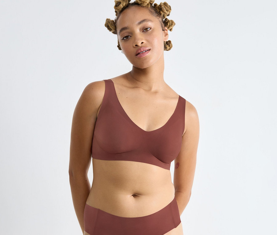 Frau mit hellbrauner Haut und Dreadlocks trägt einen braunen sloggi ZERO Feel 2.0 Bralette und passende Unterhose vor weißem Hintergrund.