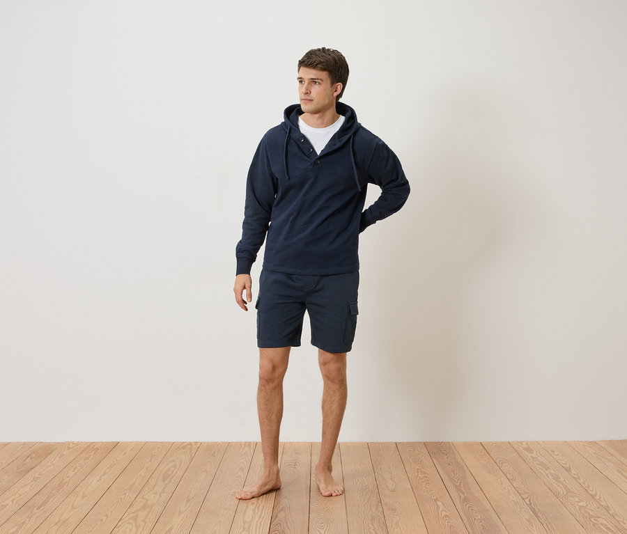 Ein Mann steht barfuß vor einer weißen Wand und trägt einen marineblauen Hoodie und Shorts.