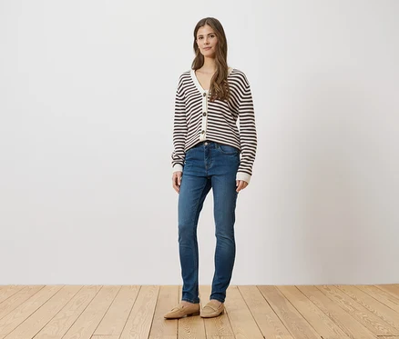 Frau in blauen Jeans, gestreifter Strickjacke und beigen Schuhen steht vor einer weißen Wand auf einem Holzboden.