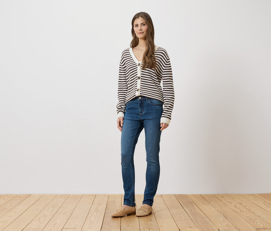 Frau in blauen Jeans, gestreifter Strickjacke und beigen Schuhen steht vor einer weißen Wand auf einem Holzboden.