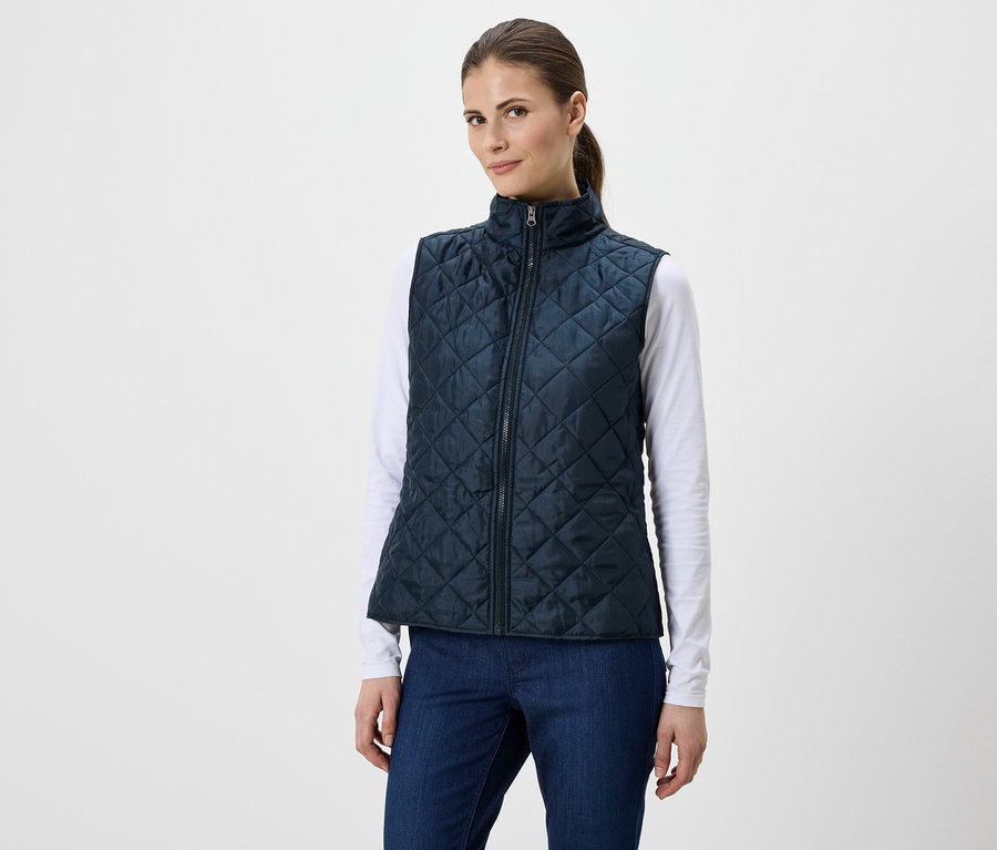 Frau mit Pferdeschwanz trägt eine marineblaue, gesteppte Weste über einem weißen Langarmshirt und blauen Jeans.