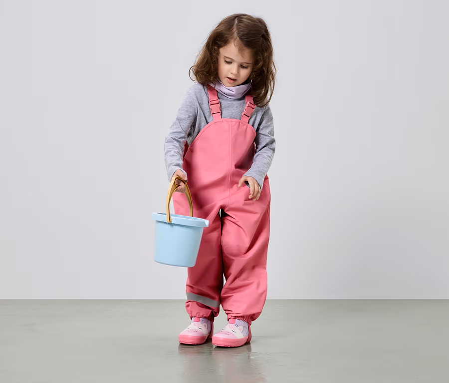 Ein Mädchen mit braunen Locken hält einen hellblauen Eimer und trägt eine rosa Latzhose und rosa Schuhe.