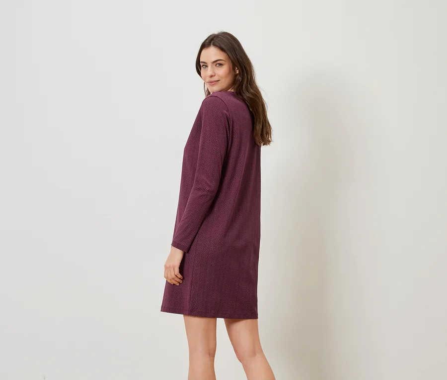 Eine Frau blickt über ihre Schulter und trägt ein burgunderrotes, langärmeliges Kleid vor einem schlichten, hellen Hintergrund.
