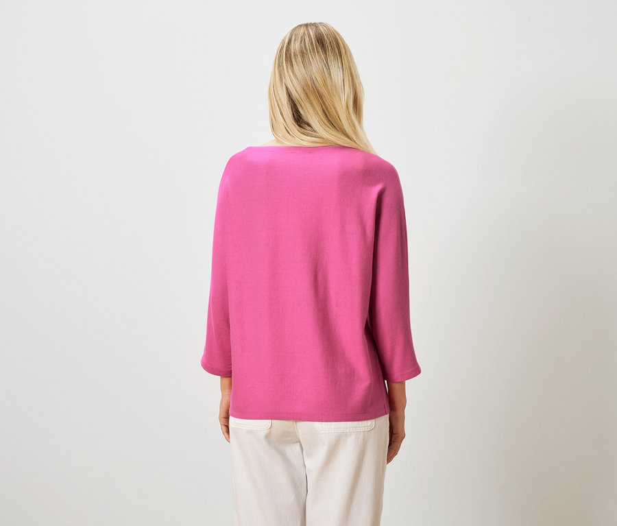 Rückansicht einer blonden Frau mit einem pinkfarbenen Oberteil und hellen Hosen vor einem weißen Hintergrund.