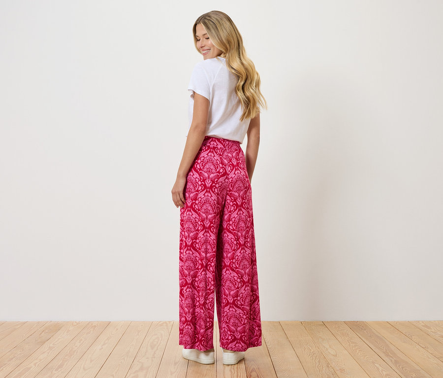 Eine Frau mit blonden Haaren trägt ein weißes T-Shirt und eine rosafarbene, weite Hose mit Muster.