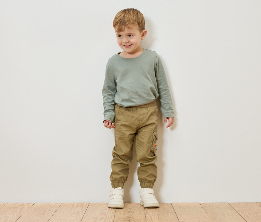 Ein Junge mit blonden Haaren steht vor einer weißen Wand und trägt ein grünes Langarmshirt, Khaki-Hosen und weiße Schuhe.