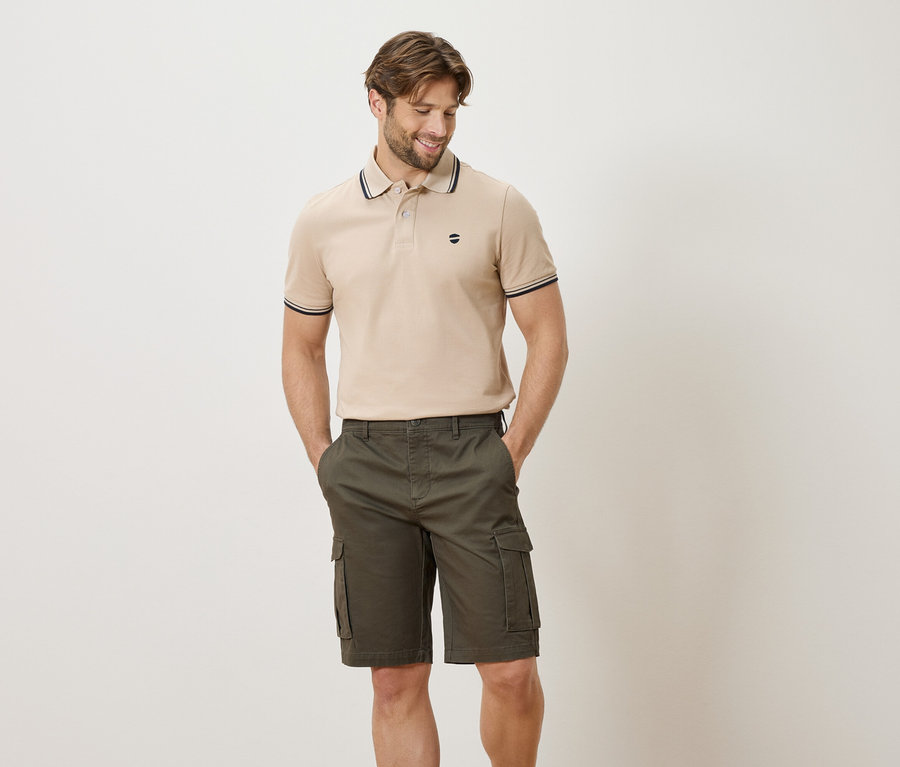 Mann mit beigefarbenem Poloshirt und olivgrünen Cargoshorts posiert mit Händen in den Hosentaschen vor einem weißen Hintergrund.