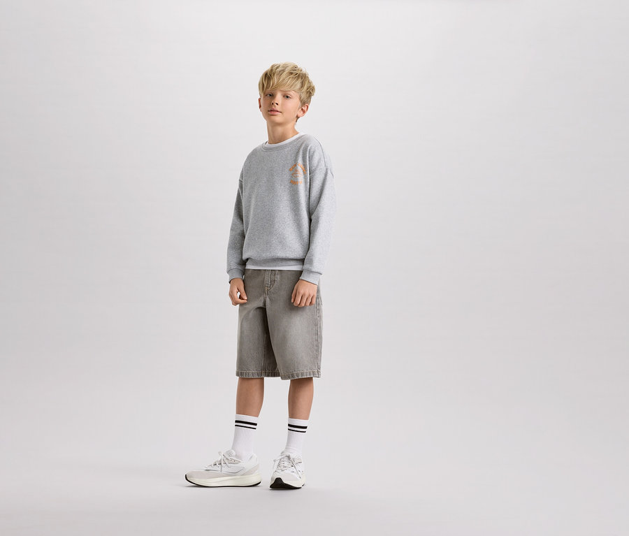 Ein blonder Junge steht in einem grauen Pullover, Shorts und weißen Socken mit Streifen.