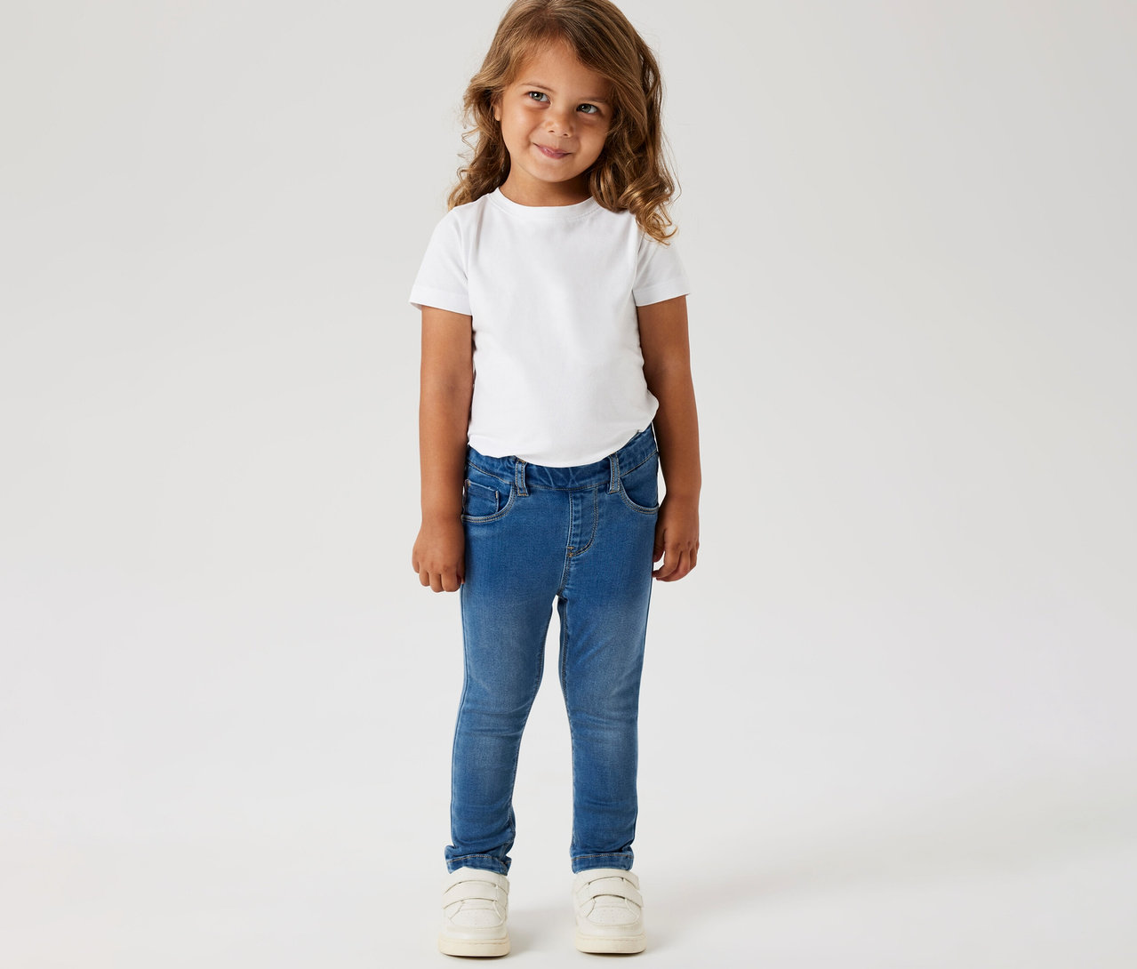 Mädchen trägt ein weißes T-Shirt und NAME IT Kinder-Jeggings und weiße Schuhe.