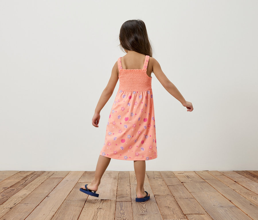 Ein Mädchen von hinten, trägt ein lachsfarbenes Sommerkleid mit Sternen und Muscheln, dazu blaue Flip-Flops, auf einem Holzboden vor einer weißen Wand.