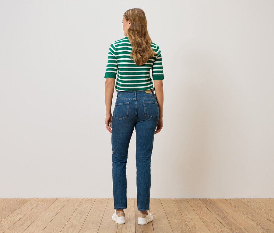 Frau von hinten in grün-weiß gestreiftem Oberteil und Jeans, steht vor einer Wand.