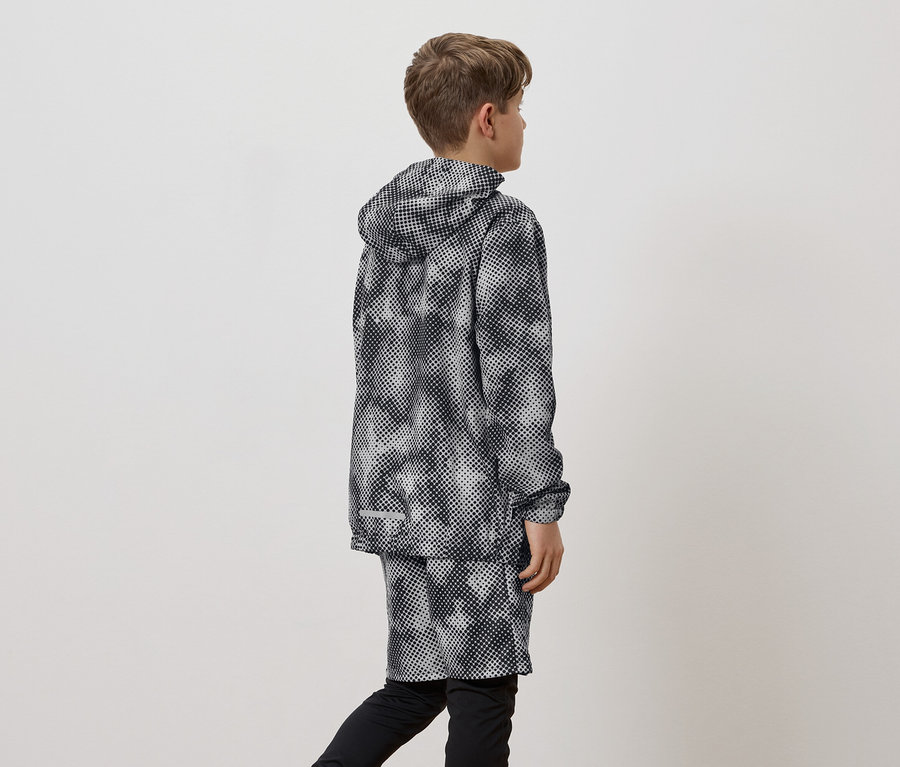 Ein Junge von hinten betrachtet, trägt eine schwarz-weiß gemusterte Regenjacke mit Kapuze und Shorts über schwarzen Leggings.
