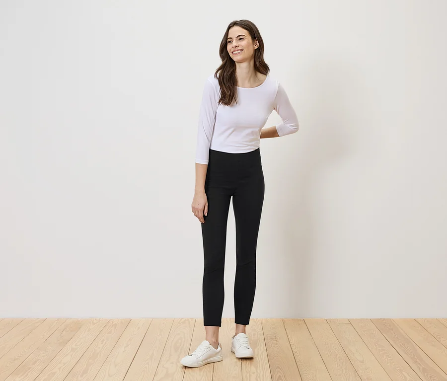 Eine Frau mit langen, braunen Haaren steht vor einer weißen Wand und trägt ein weißes Oberteil, schwarze Leggings und weiße Turnschuhe.