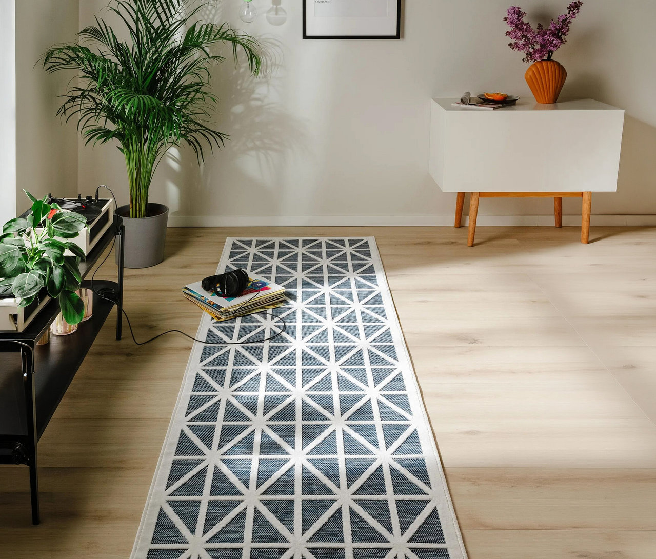 Ein Raum mit hellem Holzboden und weißen Wänden zeigt einen blau-weißen geometrischen Läufer. Links steht ein schwarzes Regal mit Plattenspieler und Pflanze, dahinter eine große Palme. Auf dem Teppich liegen Kopfhörer auf einem Stapel Schallplatten. Rechts steht ein weißer Schrank mit einer orangefarbenen Vase und lila Blumen.