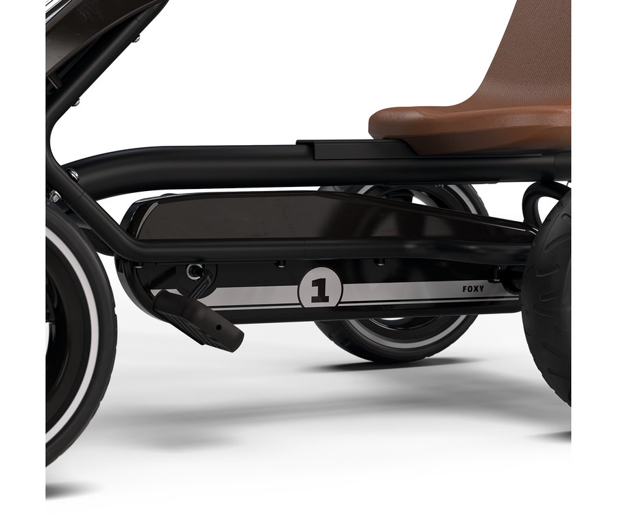 Nahaufnahme eines schwarzen Tret-Gokarts mit braunem Sitz, schwarzen Pedalen und Rädern mit weißen Seitenwänden. Ein silberner Streifen an der Seitenverkleidung zeigt einen weißen Kreis mit der Nummer '1' und den Text 'FOXY'.
