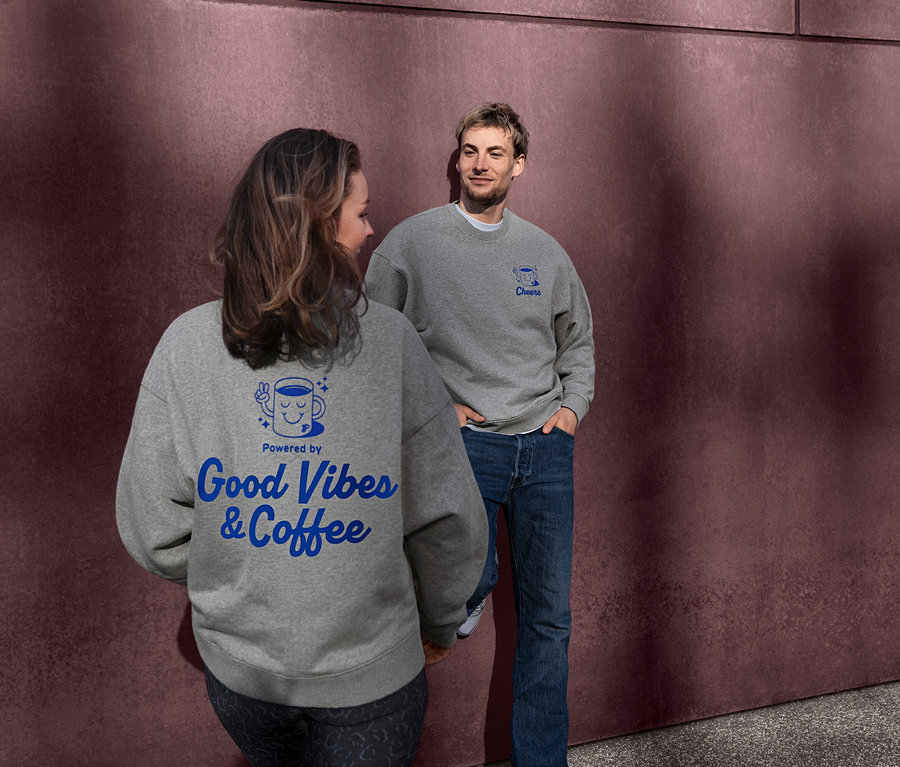 Frau und Mann tragen graue Unisex Coffee Crew Sweatshirts. Der Rücken des Sweatshirts der Frau zeigt ein blaues "Powered by Good Vibes & Coffee" Design. Der Mann blickt die Frau an, sein Sweatshirt hat ein blaues "Cheers" Design.