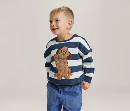 Ein kleiner Junge steht lächelnd und blickt nach oben. Er trägt ein weiß-blau gestreiftes Sweatshirt mit einer braunen Hundeapplikation und eine blaue Cordhose.