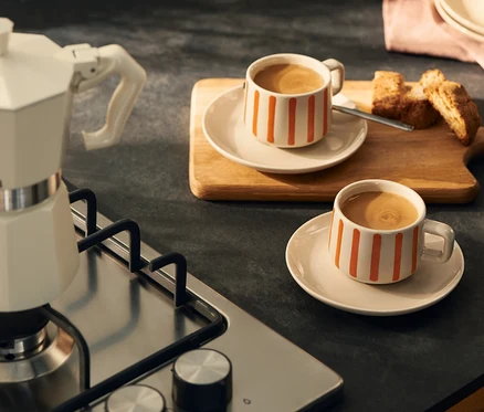 Eine cremefarbene Mokkakanne steht auf einem Gaskochfeld neben zwei weißen Tassen mit orangefarbenen Streifen, gefüllt mit Kaffee, auf einer dunklen Küchenarbeitsplatte. Biscotti liegen auf einem Holzbrett hinter einer Tasse.