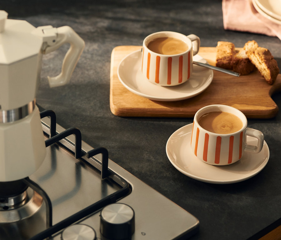 Eine cremefarbene Mokkakanne steht auf einem Gaskochfeld neben zwei weißen Tassen mit orangefarbenen Streifen, gefüllt mit Kaffee, auf einer dunklen Küchenarbeitsplatte. Biscotti liegen auf einem Holzbrett hinter einer Tasse.