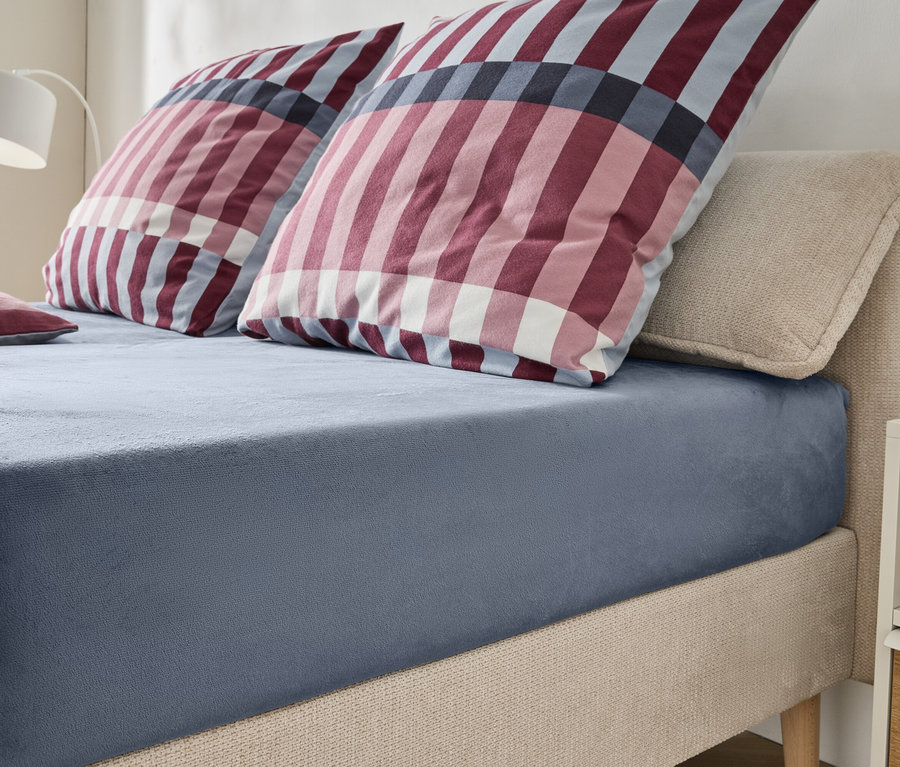 Ein Bett mit beigem, gepolstertem Kopfteil und Rahmen, bezogen mit einem blaugrauen Spannbetttuch. Zwei große Kissen und ein kleines Kissen mit rot-weiß-blauem Karomuster liegen auf dem Bett. Eine weiße Lampe und ein Nachttisch sind im Hintergrund sichtbar.