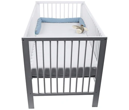 Weißes und graues Babybett mit weißer Matratze, einer blau gemusterten Bettschlange an einer Seite und einem kleinen Plüsch-Esel am Kopfende.