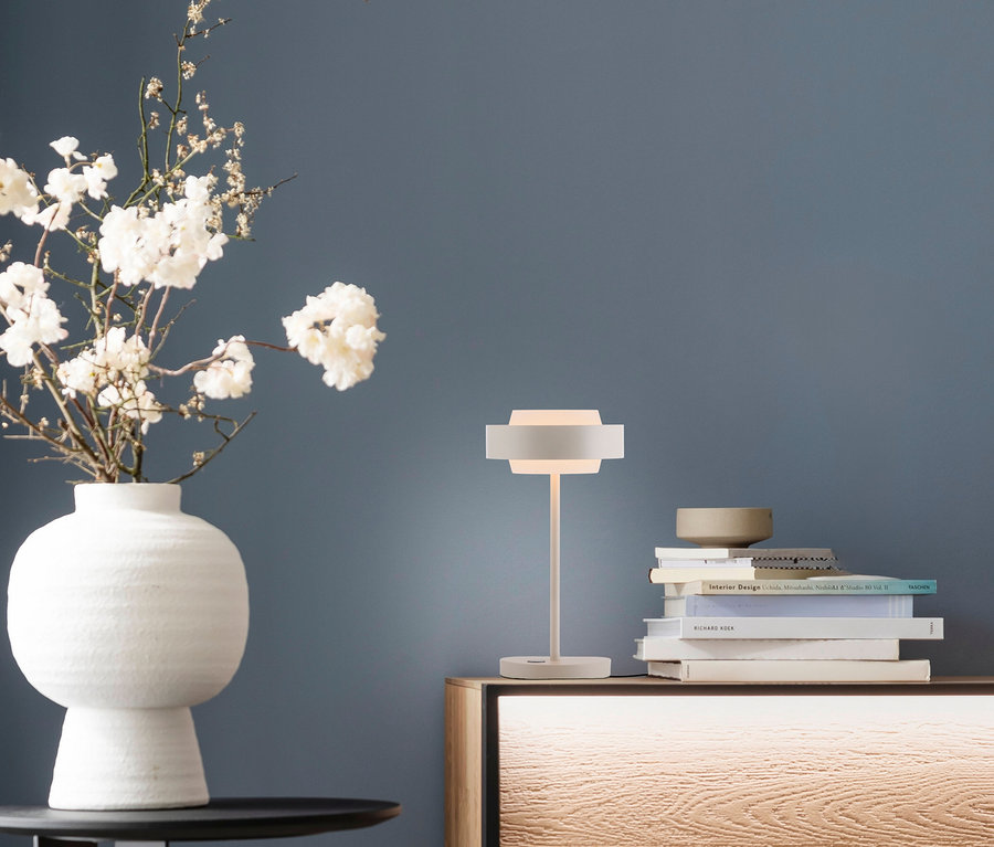 Eine beleuchtete weiße Tischlampe steht auf einem Holzschrank neben einem Stapel Bücher und einer kleinen Schale. Links davon steht eine weiße Vase mit weißen Blumen auf einem schwarzen Tisch vor einer dunkelblauen Wand.
